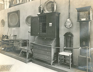 Furniture on display behind rope, including chairs, table, tall writing desk, and grandfather clock.